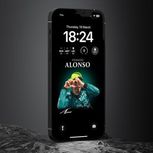 Pode incluir: Smartphone preto exibindo uma tela de bloqueio com uma foto de Fernando Alonso, a hora 18:24 e a data quinta-feira, 19 de março. O telefone está sobre uma superfície escura e texturizada.