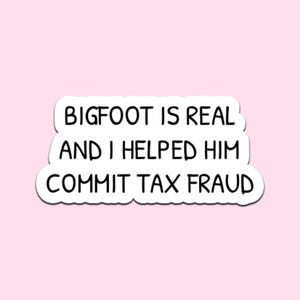 May include: A white sticker with black text on a light pink background. The text reads "BIGFOOT IS REAL AND I HELPED HIM COMMIT TAX FRAUD" in a playful, handwritten style. The sticker has a white border.