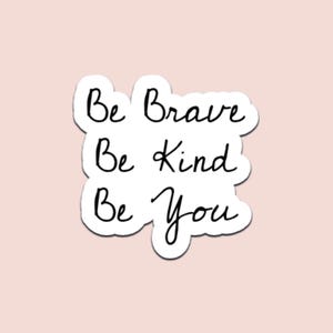 Op de afbeelding: Een witte sticker met de handgeschreven tekst "Be Brave, Be Kind, Be You" in zwarte letters. De sticker heeft een witte rand en staat op een lichtroze achtergrond. De sticker is een motiverend item.