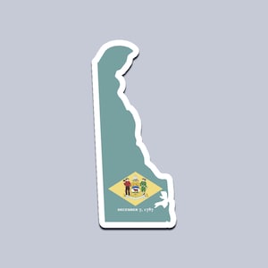 May include: A sticker in the shape of the state of Delaware, colored teal with a white border. The state seal is in the lower center, with the text "DECEMBER 7, 1787" below it. The sticker is on a gray background.