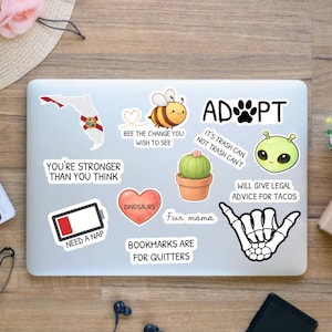 Peut inclure: Un ordinateur portable recouvert d'autocollants avec divers motifs et textes. Les autocollants incluent un contour de la Floride, une abeille, un cactus, un c&oelig;ur, un extraterrestre et une main de squelette. Le texte comprend "ADOPT" et "Need a Nap".