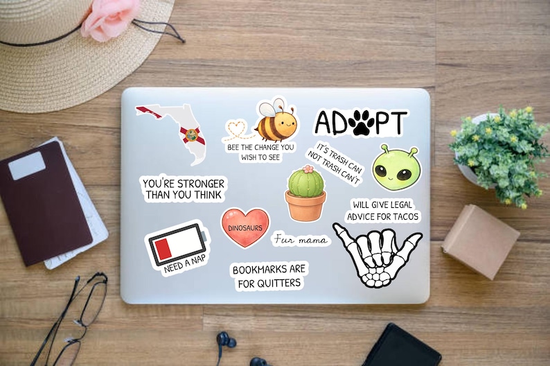 Peut inclure: Un ordinateur portable recouvert d'autocollants avec divers motifs et textes. Les autocollants incluent un contour de la Floride, une abeille, un cactus, un c&oelig;ur, un extraterrestre et une main de squelette. Le texte comprend "ADOPT" et "Need a Nap".