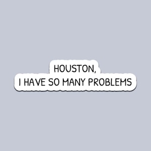Può includere: Un adesivo bianco con la frase "HOUSTON, I HAVE SO MANY PROBLEMS" in nero, con carattere sans-serif. Il testo è racchiuso in un bordo bianco di forma irregolare, su sfondo grigio chiaro.