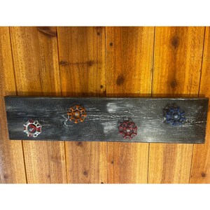 May include: A rectangular wall-mounted rack featuring a distressed black wooden board with a visible grain. Four metal valve handles in red, orange, dark red, and blue are attached to the board, serving as hooks for hanging various items.