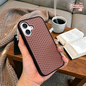May include: A mauve phone case with a black border and a textured, diamond-patterned design. The case is held in a hand, with a coffee cup and open book visible in the background. The case has a dual-lens camera cutout.