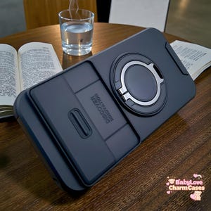 Puede incluir: Una funda de teléfono gris oscuro con un soporte de anillo incorporado y una cubierta deslizante. La funda presenta el texto "DISCOVER INNOVATION" y está sobre una superficie de madera. También se ven un vaso de agua y un libro abierto.