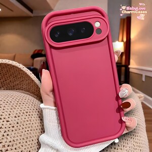 Puede incluir: Una funda de teléfono magenta con un recorte para la cámara y el texto "BabyLove CharmCases". La funda tiene un acabado liso y un borde elevado alrededor de la cámara. La funda está diseñada para proteger el teléfono.