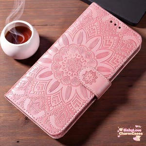 Puede incluir: Funda de teléfono rosa con un diseño floral de mandala en relieve. La funda tiene un cierre magnético y una superficie texturizada. Una taza de té está al lado de la funda, con el texto "BabyLove CharmCases".