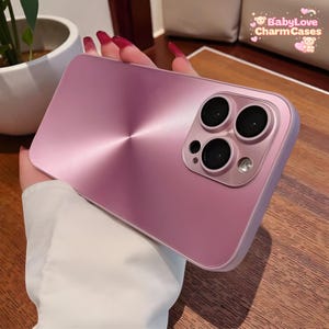 May include: A pink phone case with a brushed metal texture and a camera module featuring three lenses and a small sensor. The case has rounded edges and a smooth finish. The BabyLove Charm Cases logo is visible in the background.