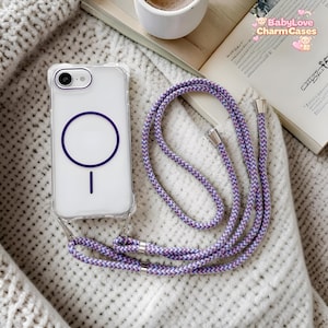Puede incluir: Funda transparente para teléfono con un diseño de círculo y línea morados, sujeta a un cordón trenzado lavanda y blanco. La funda del teléfono tiene una cámara y está sobre una superficie blanca.