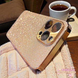 Könnte beinhalten: Eine roségoldene Glitzer-Handyhülle mit einem goldenen Kameraschutz. Die Hülle liegt auf einem rosa Handtuch. Eine weiße Teetasse und ein Buch befinden sich im Hintergrund. Der Text "BabyLove Charm Cases" ist sichtbar.