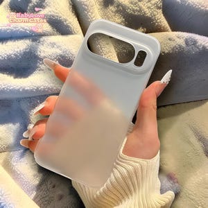 Puede incluir: Una funda de teléfono blanca translúcida con recortes para la cámara y los botones. La funda se sostiene en una mano con uñas largas y cuidadas. El texto "BabyLove Charm Cases" es visible en la esquina superior izquierda.