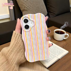 Puede incluir: Funda de teléfono con rayas pastel en tonos rosa, amarillo, azul y beige. La funda tiene un borde ondulado y recortes para la cámara del teléfono. El texto "BabyLove Charm Cases" es visible en la esquina superior izquierda.