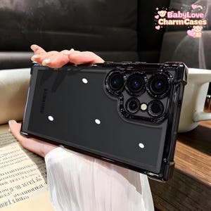 Puede incluir: Una funda transparente para teléfono con borde negro y lunares blancos. La funda tiene un logotipo de Samsung y un recorte para la cámara. La funda la sostiene una persona.