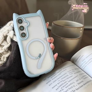 Könnte beinhalten: Eine hellblaue Handyhülle mit Katzenohren und einem Schwanzdesign. Die Hülle hat eine transparente Rückseite und einen hellblauen Rahmen. Die Handyhülle hat drei Kameraausschnitte. Der Text "BabyLove CharmCases" ist oben rechts sichtbar.