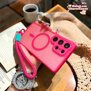 Puede incluir: Una funda de teléfono rosa brillante con una correa rosa a juego y un acento turquesa. La funda tiene un diseño circular y un módulo de cámara con múltiples lentes. El texto "BabyLove CharmCases" es visible.