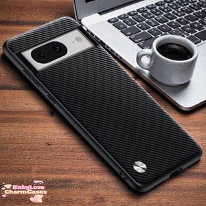 Puede incluir: Funda de teléfono negra con un patrón de fibra de carbono texturizado. La funda tiene un recorte para la cámara y un botón plateado. Un portátil y una taza de café están en el fondo. Para Google Pixel 7.