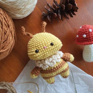 May include: A crocheted bee doll with a yellow head and a striped body in shades of yellow and brown. It has small black eyes and a pink nose. A small crochet mushroom with a red cap and white spots is also visible.