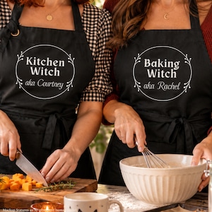 Puede incluir: Dos delantales negros con texto blanco y un borde circular de hojas. El delantal izquierdo muestra el texto Kitchen Witch aka Courtney, mientras que el derecho muestra el texto Baking Witch aka Rachel. Ambos tienen lazos en la cintura y cuello.