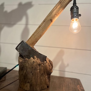 以下が含まれることがあります： 古びた木製の土台と斧の頭で作られた素朴なランプ。木製の腕が電球を支えています。 ランプはヴィンテージスタイルの電球を使用し、温かい光を放ちます。 土台は自然な質感の木材です。