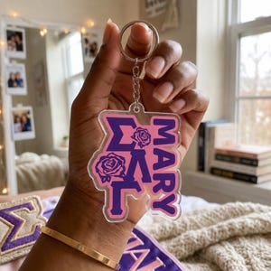 May include: A clear acrylic keychain featuring a purple and pink design with the text MARY and Greek letters. The design includes two purple rose illustrations and a silver metal split ring attached to a short chain, showcasing a layered aesthetic.