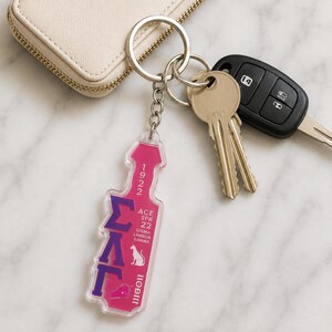 May include: A clear acrylic keychain shaped like a paddle, featuring a vibrant pink background with purple Greek letters, the text 1922, ACE SPR 22, SIGMA LAMBDA GAMMA, and IIOIII, along with a small white panther silhouette and a unicorn head icon.