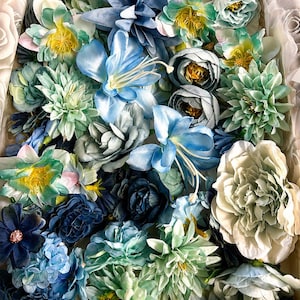 Puede incluir: Una colecci&oacute;n de flores artificiales en varios tonos de azul, verde azulado y blanco. El surtido incluye rosas, peon&iacute;as y lirios con p&eacute;talos en capas, algunos con centros amarillos o detalles de purpurina, empaquetados estrechamente en una caja.