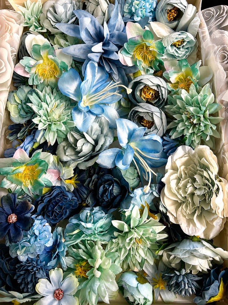 Puede incluir: Una colecci&oacute;n de flores artificiales en varios tonos de azul, verde azulado y blanco. El surtido incluye rosas, peon&iacute;as y lirios con p&eacute;talos en capas, algunos con centros amarillos o detalles de purpurina, empaquetados estrechamente en una caja.