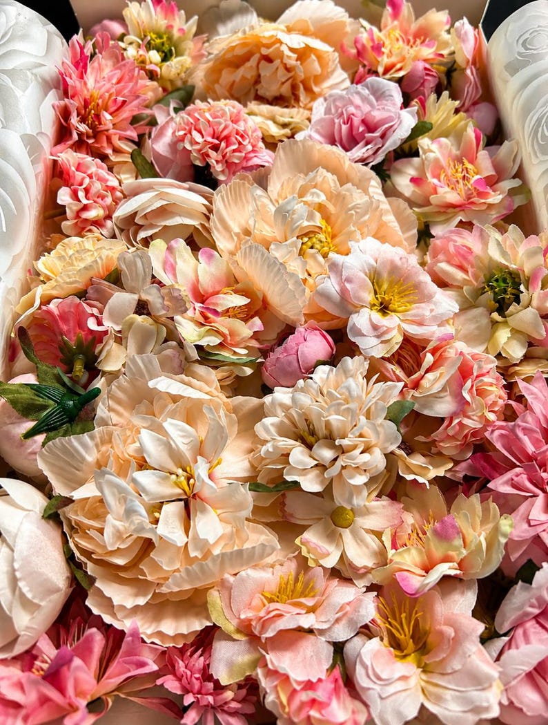 Puede incluir: Una colecci&oacute;n de flores artificiales en tonos melocot&oacute;n, rosa suave y crema. El arreglo presenta varios tipos de flores, incluyendo peon&iacute;as, dalias y flores m&aacute;s peque&ntilde;as con p&eacute;talos en capas, tallos verdes y centros amarillos.