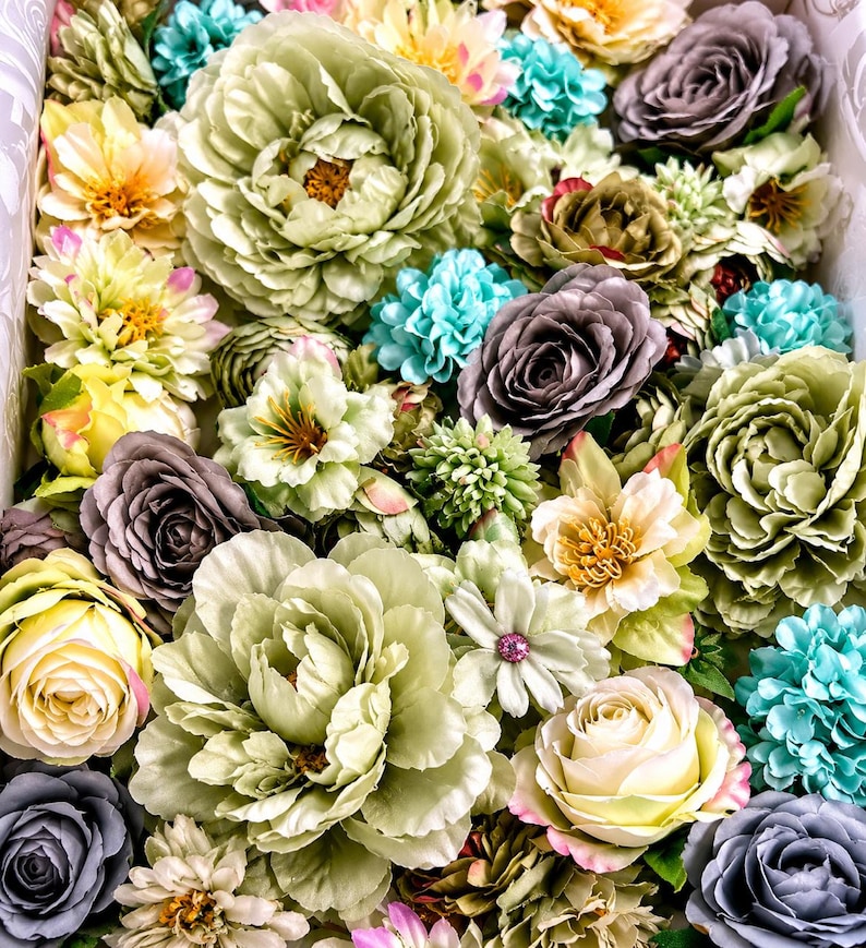 Puede incluir: Una colecci&oacute;n de flores artificiales en varios tonos de verde salvia, morado polvoriento, crema y turquesa brillante. El arreglo presenta p&eacute;talos en capas de rosas, peon&iacute;as y peque&ntilde;as flores de relleno, creando una exhibici&oacute;n floral densa.