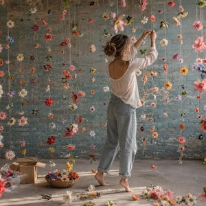 May include: A white long-sleeved top and light blue denim jeans. The scene features numerous colorful artificial flowers suspended from the ceiling on thin strings, creating a vertical floral display against a textured blue brick wall.
