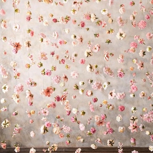 Puede incluir: Un colgante decorativo de pared que presenta numerosas flores artificiales individuales de color rosa, blanco y melocot&oacute;n suspendidas de hilos verticales finos. El arreglo floral crea un efecto de cascada sobre un fondo neutro y texturizado.