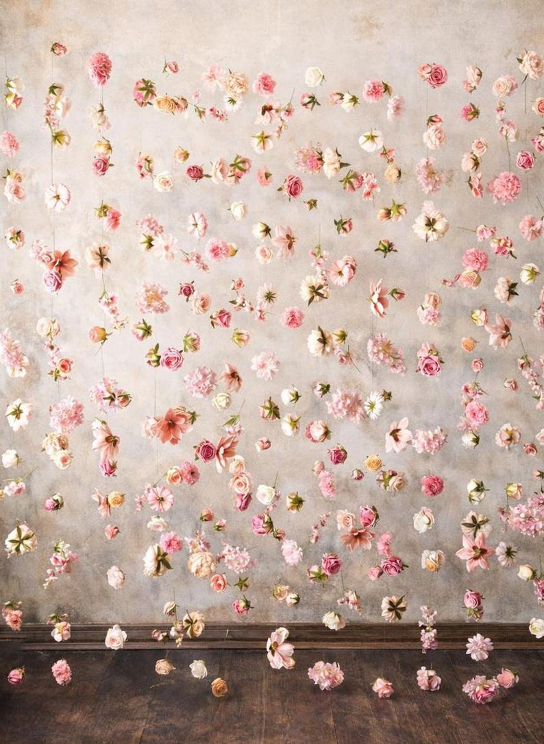 Puede incluir: Un colgante decorativo de pared que presenta numerosas flores artificiales individuales de color rosa, blanco y melocot&oacute;n suspendidas de hilos verticales finos. El arreglo floral crea un efecto de cascada sobre un fondo neutro y texturizado.