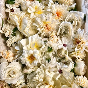 Puede incluir: Una colecci&oacute;n de flores artificiales en tonos crema, marfil y melocot&oacute;n p&aacute;lido. El arreglo presenta varias flores, incluyendo rosas, peon&iacute;as y dalias con p&eacute;talos en capas, tallos verdes y peque&ntilde;os centros oscuros en algunas flores.