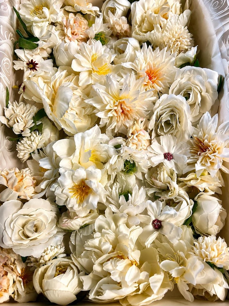 Puede incluir: Una colecci&oacute;n de flores artificiales en tonos crema, marfil y melocot&oacute;n p&aacute;lido. El arreglo presenta varias flores, incluyendo rosas, peon&iacute;as y dalias con p&eacute;talos en capas, tallos verdes y peque&ntilde;os centros oscuros en algunas flores.