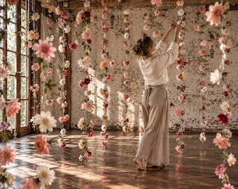 Puede incluir: Una persona con una blusa blanca de manga larga y pantalones beige holgados se encuentra en una habitación llena de numerosas cuerdas verticales de flores rosas, blancas y rojas que cuelgan desde el techo hasta el suelo de madera.