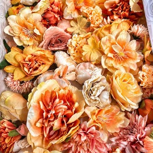 Puede incluir: Una colecci&oacute;n de flores artificiales en varios tonos de naranja, melocot&oacute;n y crema. El arreglo presenta m&uacute;ltiples tipos de flores, incluyendo rosas, peon&iacute;as y dalias, con p&eacute;talos en capas y follaje verde intercalado entre las flores.