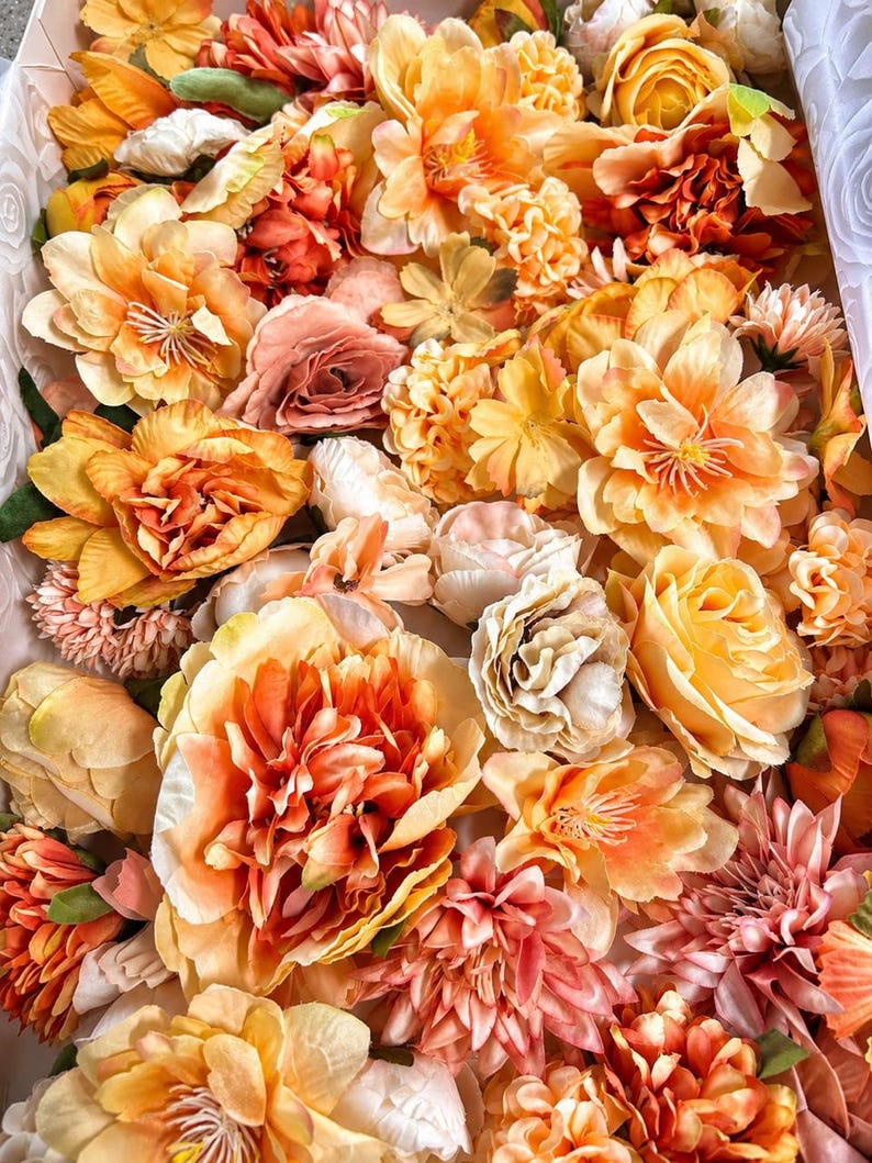 Puede incluir: Una colecci&oacute;n de flores artificiales en varios tonos de naranja, melocot&oacute;n y crema. El arreglo presenta m&uacute;ltiples tipos de flores, incluyendo rosas, peon&iacute;as y dalias, con p&eacute;talos en capas y follaje verde intercalado entre las flores.
