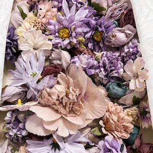 Puede incluir: Una colecci&oacute;n de flores artificiales en tonos lavanda, malva, rosa empolvado y crema. El arreglo presenta diversas flores, incluyendo peon&iacute;as, margaritas y peque&ntilde;as flores agrupadas, dispuestas de forma compacta con acentos de follaje verde.