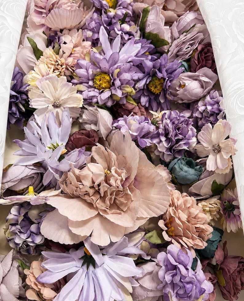 Puede incluir: Una colecci&oacute;n de flores artificiales en tonos lavanda, malva, rosa empolvado y crema. El arreglo presenta diversas flores, incluyendo peon&iacute;as, margaritas y peque&ntilde;as flores agrupadas, dispuestas de forma compacta con acentos de follaje verde.