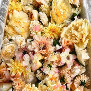 Puede incluir: Una colecci&oacute;n de flores artificiales en tonos amarillo, crema y melocot&oacute;n suave, empaquetadas estrechamente en una caja rectangular. El surtido incluye varios tipos de flores como peon&iacute;as, dalias y rosas con p&eacute;talos de tela.