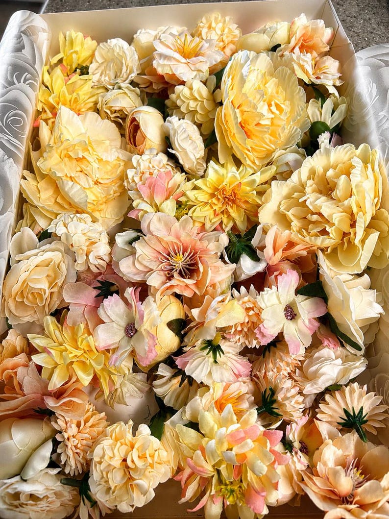 Puede incluir: Una colecci&oacute;n de flores artificiales en tonos amarillo, crema y melocot&oacute;n suave, empaquetadas estrechamente en una caja rectangular. El surtido incluye varios tipos de flores como peon&iacute;as, dalias y rosas con p&eacute;talos de tela.