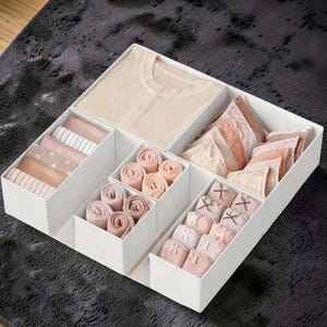 May include: A white compartmentalized storage box filled with folded and rolled undergarments and clothing. The items are in shades of beige, pink, and white. The box is on a dark gray surface.