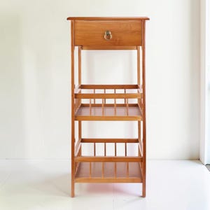 May include: A tall, wooden shelf unit with a drawer and three open shelves. The unit is made of light brown wood and features a brass-colored drawer pull. The shelves have a slatted design, and the unit stands on four legs.