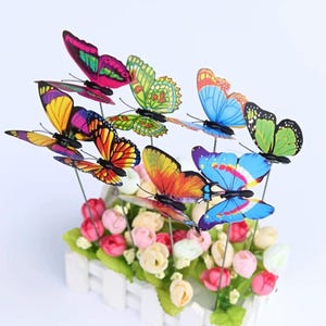 Może przedstawiać: Kolekcja kolorowych dekoracji w kształcie motyli na metalowych prętach. Motyle mają żywe odcienie różu, żółci, pomarańczy, błękitu i zieleni. Ułożone są nad grządką sztucznych kwiatów w białej donicy z płotkiem.