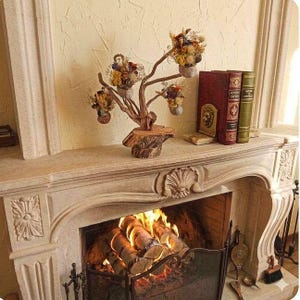 Puede incluir: Una repisa decorativa de chimenea con una escultura de árbol de madera que sostiene pequeños arreglos florales. Dos libros antiguos y un sujetalibros de león se encuentran a la derecha del árbol. La chimenea de abajo tiene un fuego ardiendo con leños.