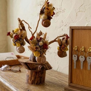 Puede incluir: Una escultura decorativa de madera con flores secas y detalles de conchas de caracol. La escultura tiene una base y ramas de madera natural, con pequeños ramos de flores secas de colores. Un llavero de madera está a la derecha.