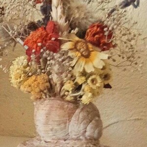 Pode incluir: Um arranjo de flores secas num vaso feito de material tecido. O buquê apresenta uma variedade de flores secas em tons de amarelo, laranja, vermelho e castanho. O vaso tem uma base redonda e uma superfície texturizada.
