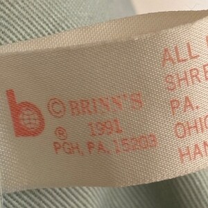 Puede incluir: Una etiqueta de tela blanca con texto rosa. La etiqueta dice "b &copy;BRINN'S @1991 PGH, PA, 15203 ALL NEW SHREAR PA. RE OHIO HAND".