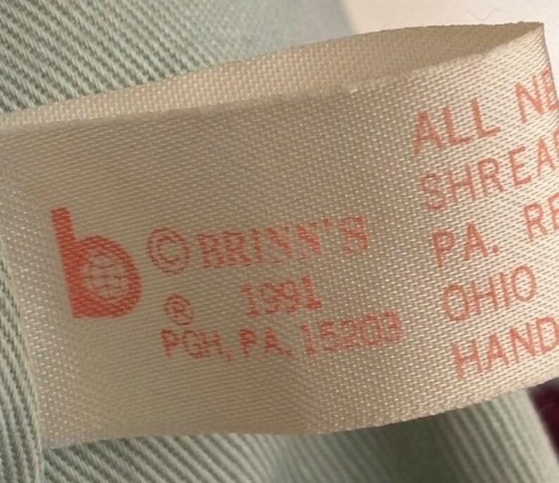 Puede incluir: Una etiqueta de tela blanca con texto rosa. La etiqueta dice "b &copy;BRINN'S @1991 PGH, PA, 15203 ALL NEW SHREAR PA. RE OHIO HAND".